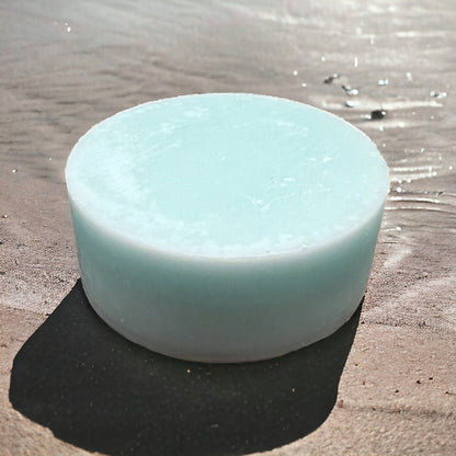 a light blue round and smooth conditioner bar sits on a wet sandy beach with the tide coming up behind it. 