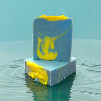 two bars of handmade soap, one sitting on top of the other that is sitting on its side to showcase the top design of the bars. with swirls of neon yellow and blue, resembling a melted state, on a reflective rippling splash blue surface against a blue background.