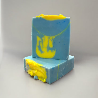 two bars of handmade soap, one sitting on top of the other that is sitting on its side to showcase the top design of the bars. with swirls of neon yellow and blue, resembling a melted state sitting on a grey surface against a grey background