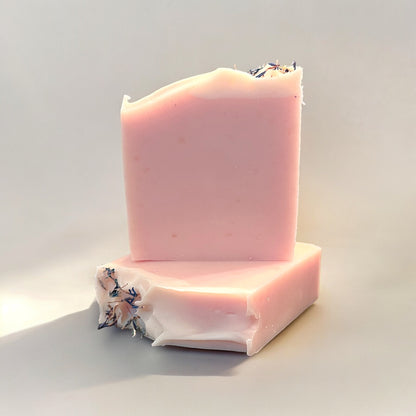 Two bars of handmade soap with a light pink color and a white top design, featuring dried blue cornflower petals as garnished on top of each bar. the bars sit on a white surface against a white background