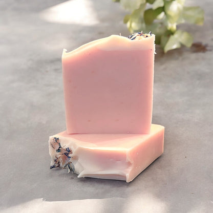 Two bars of handmade soap with a light pink color and a white top design, featuring dried blue cornflower petals as garnished on top of each bar. the bars are sitting on a grey surface with a blurred background that has a houseplant in the background.