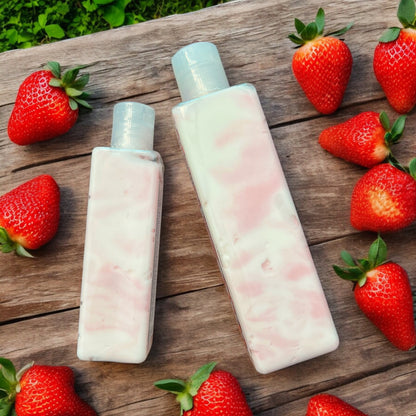 Two bottles of Strawberries & Cream Body Creme Lotion laying on a flat surface and fresh strawberries scattered around the bottles. there is grass in the background. The bottles are clear and the lotion inside is swirls of red and white. 