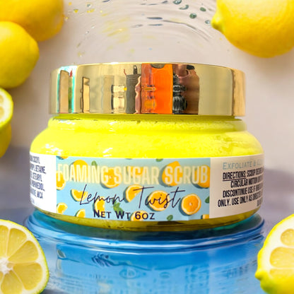 A jar of Lemon Twist Foaming Sugar Scrub by Dandy Moon Soapery, with lemon and orange design elements on the label, indicating a zesty lemon scent. the jar is clear and the foaming sugar scrub is bright yellow and the lid of the jar is gold. the jar sits on a reflective blue surface against a white background. there are lemons scattered around the jar. 