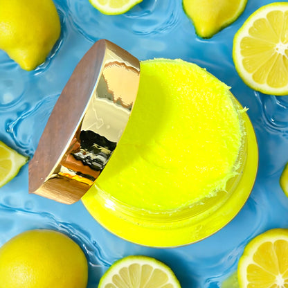 an open jar of foaming sugar scrub is seen from the top looking down into the jar. the lid of the jar is gold and is laying on the side of the jar. the bright yellow foaming sugar scrub is visible. the jar sits on a reflective bright blue surface with lemons scattered around the jar. 