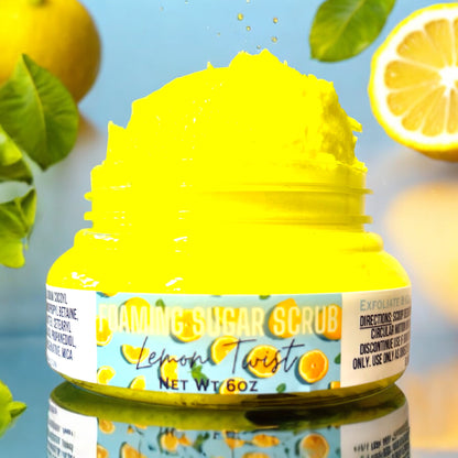 A jar of Lemon Twist Foaming Sugar Scrub by Dandy Moon Soapery, with lemon and orange design elements on the label, indicating a zesty lemon scent. the jar is clear and the foaming sugar scrub is bright yellow and heaping out of the open jar. the jar sits on a reflective surface against a blue background. there are lemons and green leaves falling around the jar in the background. 