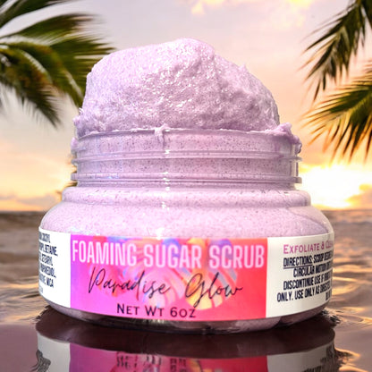 A jar of Paradise Glow Foaming Sugar Scrub by Dandy Moon Soapery, featuring a purple label and foamy purple scrub visible through the jar, and heaped over the top of the jar that is open set against a tropical background. it sits on a reflective ripple surface. there are palm trees in the background.