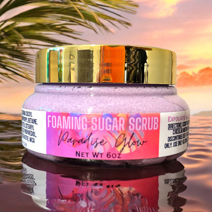 a jar of foaming sugar scrub in the paradise glow scent. the jar is clear with a gold top. the foaming sugar scrub inside it is purple. the label is bright pink with purple palm tree leaves. the jar sits on a reflective ripple surface and a sunset with palm trees are in the background. 