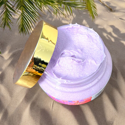 a jar of foaming sugar scrub is seen from the top looking down into the jar. the scrub is purple in color. the gold top is laid on the side of the jar. the jar sits in the sand with palm tree to the left side. the shadows from the palm tree are seen on the ground