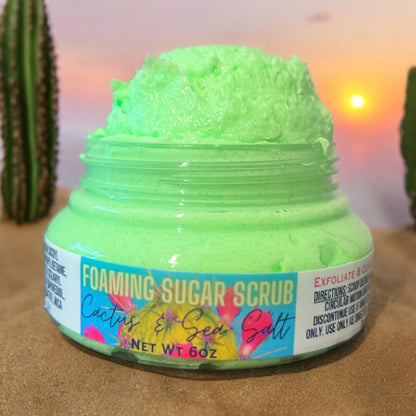 A jar of Cactus & Sea Salt Foaming Sugar Scrub by Dandy Moon Soapery, in a clear round jar with bright green scrub mounded over the top of the jar. The label is bright blue with a green cactus in the background of the label, adorned with pink cactus flowers. set against a backdrop of a cactus garden in the desert.