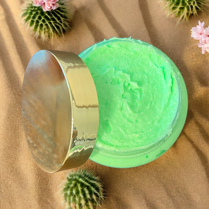 a clear jar of bright green foaming sugar scrub is seen from the top looking into the jar, the gold lid is resting on the side of the jar. the jar is sitting in desert sand with tiny cactus and cactus flowers scattered around it. 
