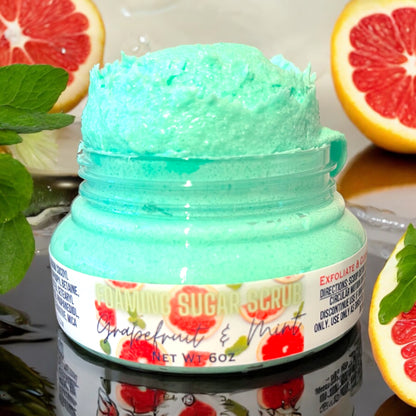 a clear jar of grapefruit and mint foaming sugar scrub sits on a reflective surface. the scrub is bright turquoise in color and spills over the top of the jar lid. the label on front is white and says foaming sugar scrub grapefruit and mint and has grapefruits on the label. the jar sits on a reflective surface and has mint leaves and fresh cut grapefruits sitting around the jar.  