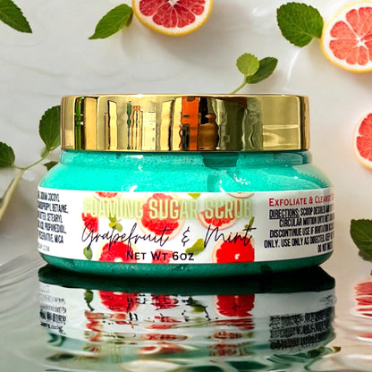 a clear jar of grapefruit and mint foaming sugar scrub sits on a reflective surface. the scrub is bright turquoise in color in the closed jar. the jar has a gold top. the label on front is white and says foaming sugar scrub grapefruit and mint and has grapefruits on the label. the jar sits on a reflective water rippled surface and has mint leaves and fresh cut grapefruits sitting around it