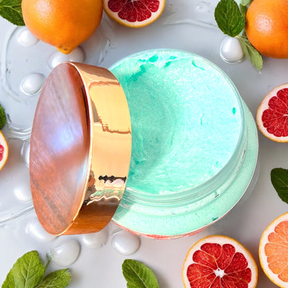 a jar of bright turquoise green is seen from the top to show into the jar. the jar is open and the gold lid sits rested on the side of the jar. the jar sits on a rippled water surface and there are mint leaves and grapefruits sitting all around the jar.  