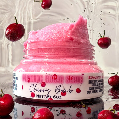 A clear jar of cherry bomb foaming sugar scrub, the scrub is heaping over the top of the open jar and the scrub is red in color. it sits on a reflective surface with water splashing and rippling around it. there are cherries falling around it and scattered around the jar. the background is water dropping and white. the label on the jar is white with cherries on it and says foaming sugar scrub cherry bomb 