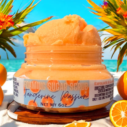 a jar or orange tangerine passion foaming sugar scrub heaped up over the rim of  the jar. the jar sits on a wooden surface on a beach, with fresh oranges scattered around it. there are exotic flowers in the background, and the ocean in the background. 