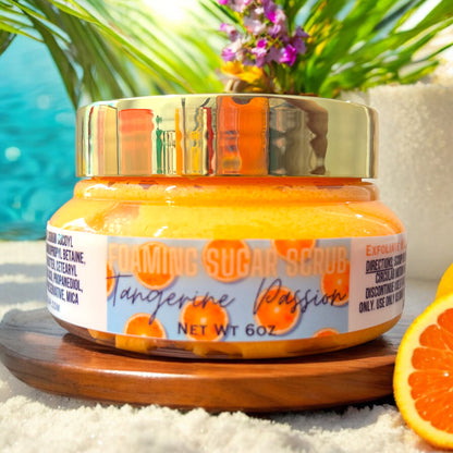 a closed clear jar of orange tangerine passion foaming sugar scrub. the jars lid is gold and the label says foaming sugar scrub and tangerine passion. the label is blue with oranges on it. the jar sits on a wooden surface, which is resting in the sand. there are fresh oranges next to the jar. there are palm trees and exotic flowers in the background