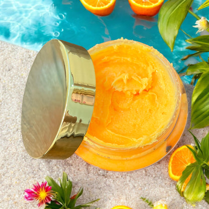shows the inside of the opened sugar scrub from the top, the gold lid to the jar rests on the side of the jar. the jar sits on sand with turquoise water coming up next to the jar in the sand. there are fresh oranges scattered in the sand and floating in the water. there are also flowers and palm leaves scattered around. 