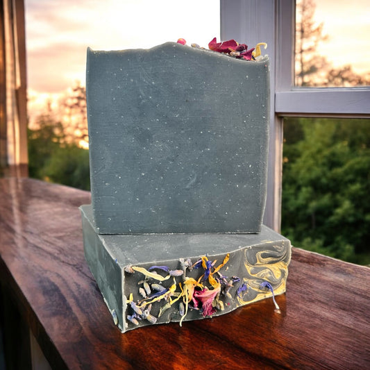 A bar of Dandy Moon Soapery's Practical Magic soap sits on top of another bar that is laying flat so the bars dried flowers are displayed. Both sit on a wooden surface with a scenic outdoor background. The soap has a deep charcoal infused hue, giving it a rich and earthy appearance. The top is adorned with dried lavender, Rose petals, blue cornflower, and Eucalyptus, adding a natural and enchanting touch. This handcrafted soap embodies the essence of nature and luxury.