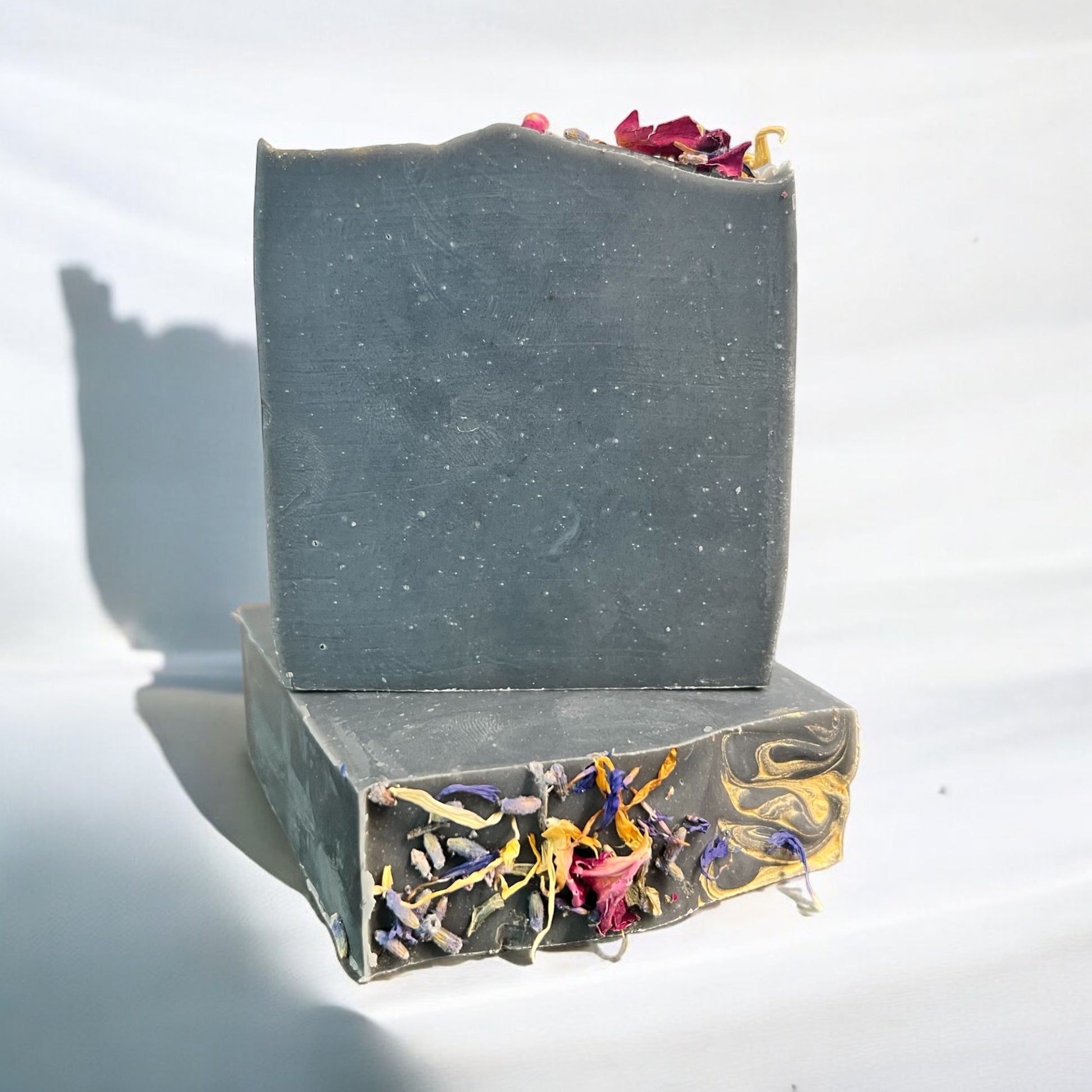 A bar of Dandy Moon Soapery's Practical Magic soap sits on top of another bar that is laying flat so the bars dried flowers are displayed. Both sit on a white surface with a white background. The soap has a deep charcoal infused hue, giving it a rich and earthy appearance. The top is adorned with dried lavender, Rose petals, blue cornflower, and Eucalyptus, adding a natural and enchanting touch. This handcrafted soap embodies the essence of nature and luxury.