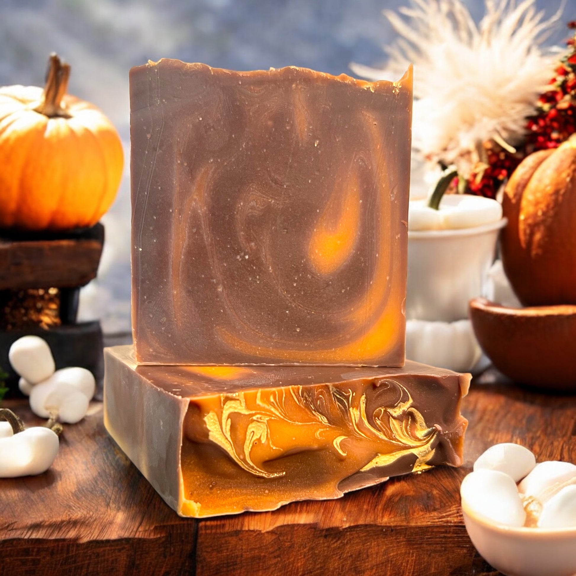 A bar of Dandy Moon Soapery's autumn inspired soap - Pumpkin S'mores bar- sits on top of another bar that is laying flat, to display the gold swirl top design. Both sit on a rustic wooden surface, surrounded by festive pumpkins, marshmallows, and cozy fall decorations. The soap has warm brown tones with swirls of golden orange, reminiscent of toasted marshmallows and seasonal warmth. The intricate marbling on the top of the bar showcases the artisanal craftsmanship. 