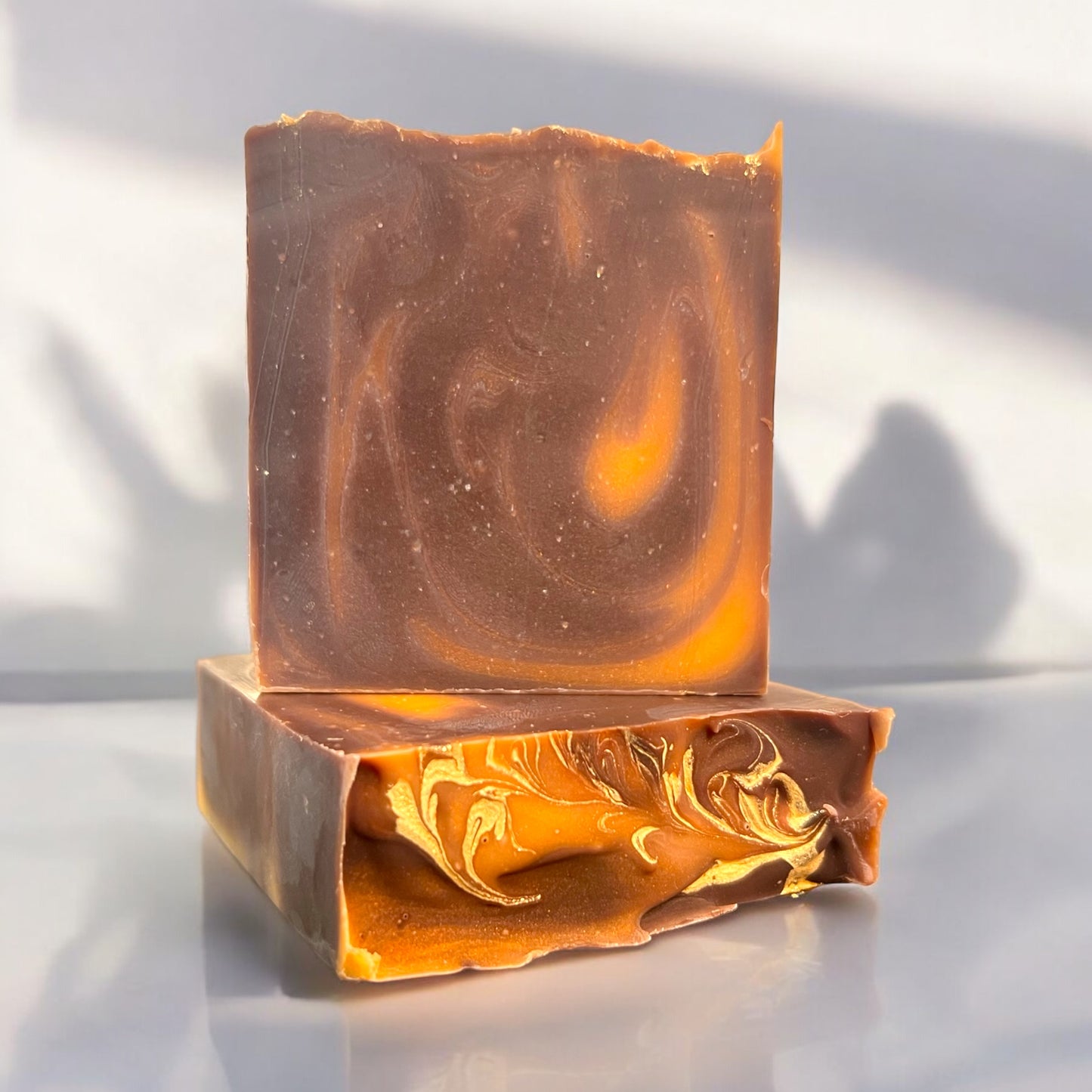 A bar of Dandy Moon Soapery's autumn inspired soap - Pumpkin S'mores bar- sits on top of another bar that is laying flat, to display the gold swirl top design. Both sit on a white surface, against a white background. The soap has warm brown tones with swirls of golden orange, reminiscent of toasted marshmallows and seasonal warmth. The intricate marbling on the top of the bar showcases the artisanal craftsmanship. 