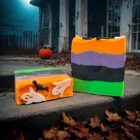 Two Bars of Dandy Moon Soapery's Spooky Season soap are displayed outdoors, set against a Halloween backdrop with fallen leaves and an old haunted looking house. The soap features vibrant layers of green, black, orange, and purple, with Halloween themed designs on top like white ghosts and black bats embedded in the top orange layer. The fruity candy scent and playful colors capture the spirit of Halloween, making this handcrafted soap a fun seasonal treat.  