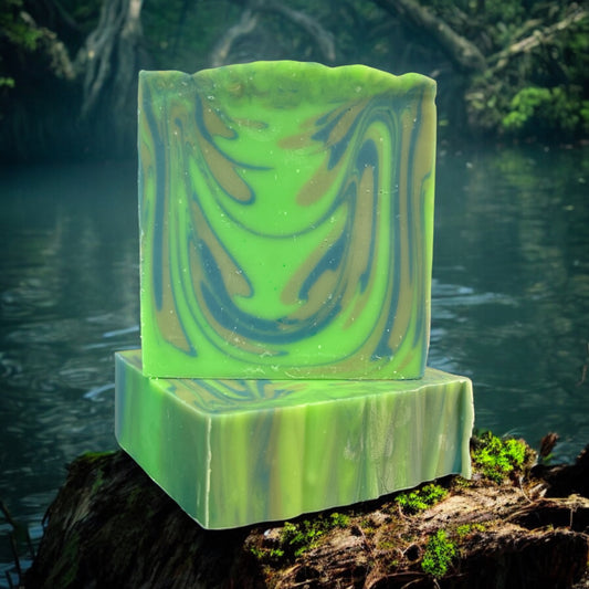 A bar of Dandy Moon Soapery's Swamp Thing soap is sitting on top of another bar that is laying flat in order to display the top lined design, and displayed outdoors with a lush swamp like background of water and greenery. The bars feature vibrant green tones with swirl patterns in earthy shades of brown and dark blue, mimicking the mysterious depths of a swamp. This handcrafted soap captures a natural, untamed aesthetic, evoking the essence of the wild and adventurous spirit of the outdoors. 