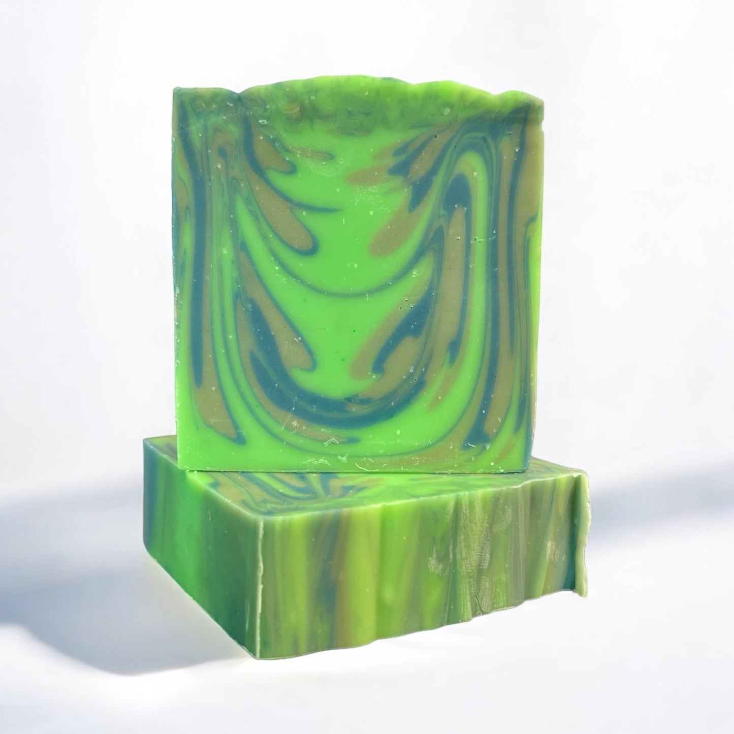 A bar of Dandy Moon Soapery's Swamp Thing soap is sitting on top of another bar that is laying flat in order to display the top lined design, and displayed on a white surface against a white background. The bars feature vibrant green tones with swirl patterns in earthy shades of brown and dark blue, mimicking the mysterious depths of a swamp. This handcrafted soap captures a natural, untamed aesthetic, evoking the essence of the wild and adventurous spirit of the outdoors. 