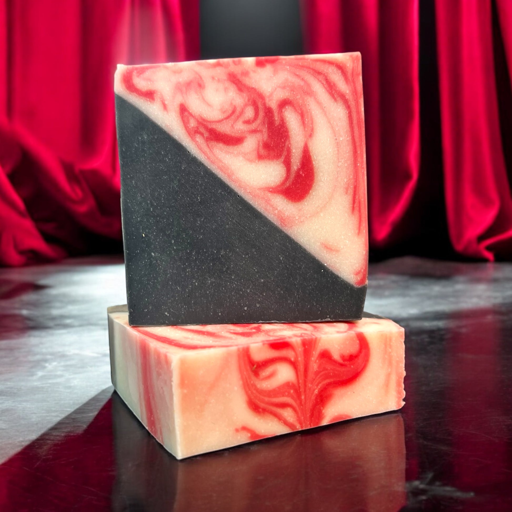 a bar of soap that is half black and half red and white swirls, sitting on top of another bar of the same design to showcase the top swirls. both bars are sitting on a state with red silk theatre curtains draped in the background. 