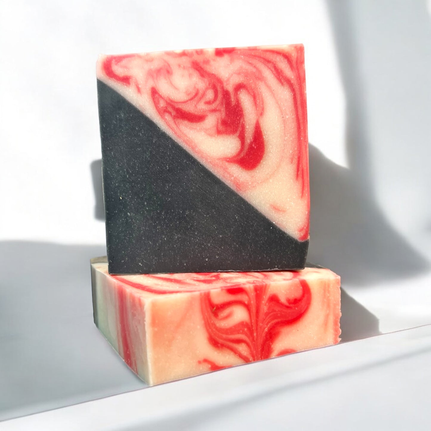 a bar of soap that is half black and half red and white swirls, sitting on top of another bar of the same design to showcase the top swirls. the background is white to showcase the bars designs. 