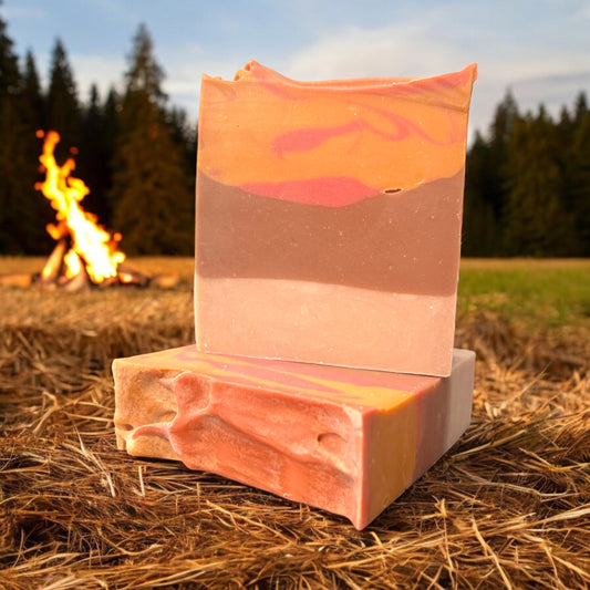 a bar of soap with gradient color scheme of tan and brown with swirls of orange and red at the top, sitting on another bar of soap in the same design. both bars are sitting in a field with a tree line in the background and a bonfire set off in the distance.  