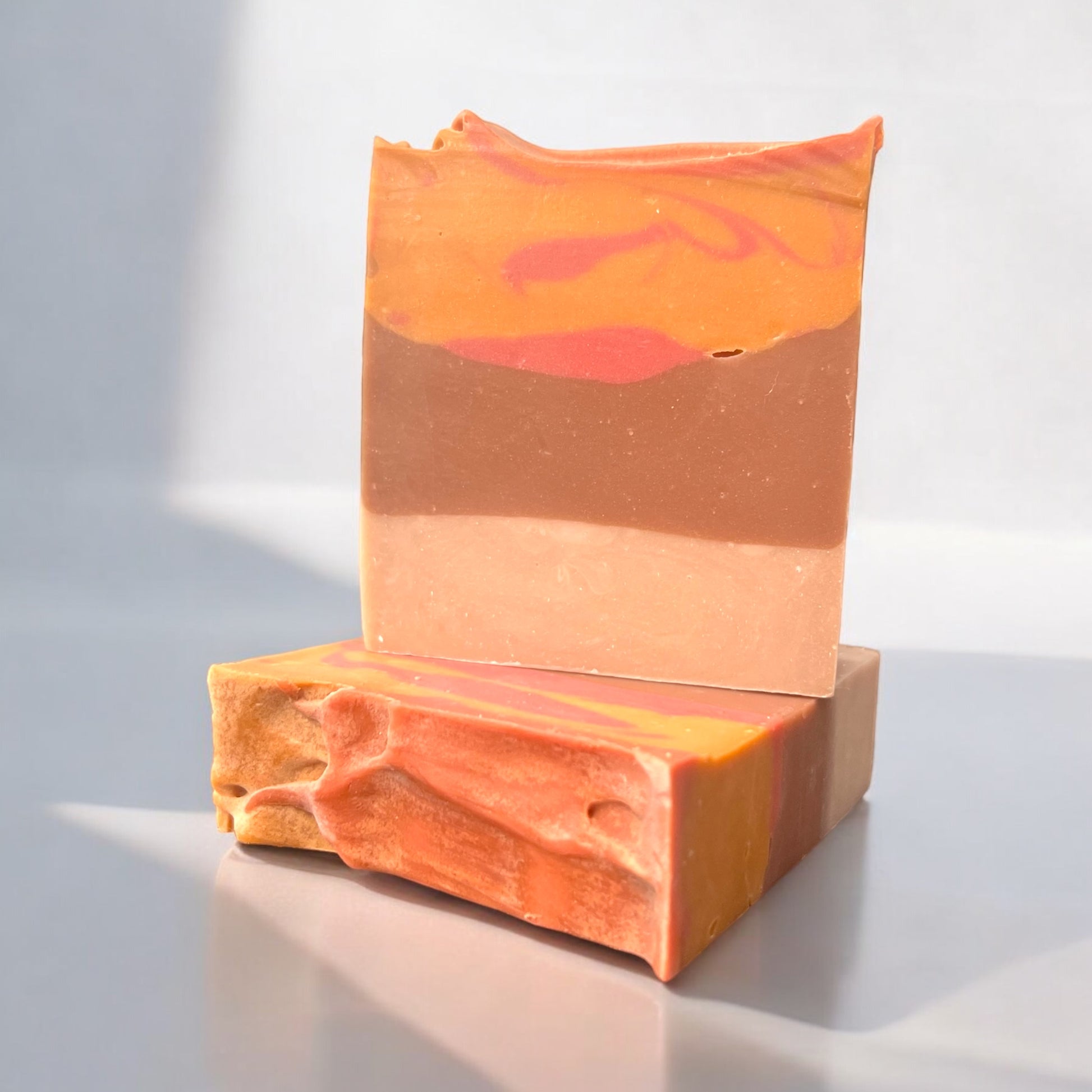 a bar of soap with gradient color scheme of tan and brown with swirls of orange and red at the top, sitting on another bar of soap in the same design. the background is white to showcase the bars alone. 