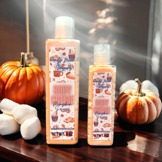 Two bottles of Dandy Moon Soapery's Pumpkin S'mores Body Creme are displayed alongside decorative pumpkins and marshmallows, evoking a cozy autumn vibe. The bottles, one 8 ounce one 4 ounce, feature a festive label with illustrations of pumpkins, s'mores, and seasonal motifs. The label reads Body Creme - Pumpkin S'mores. This handcrafted body creme embodies seasonal indulgence and luxury by Dandy Moon Soapery. 