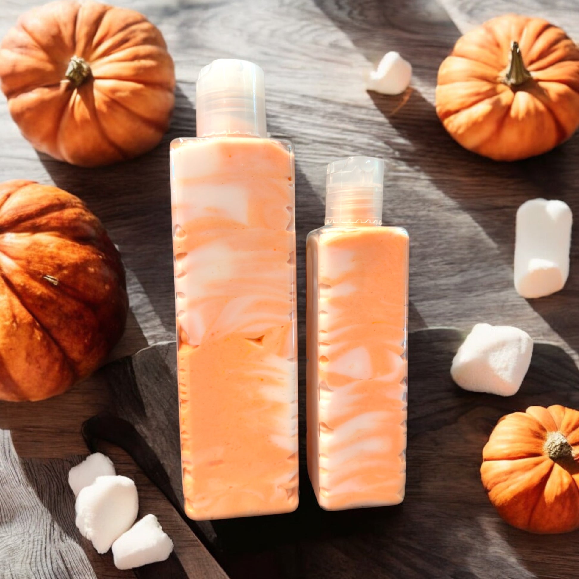 Two bottles of Dandy Moon Soapery's Pumpkin S'mores Body Creme are displayed laying on a rustic surface alongside decorative pumpkins and marshmallows, evoking a cozy autumn vibe. The bottles, one 8 ounce one 4 ounce, are laid on their side to display the lotion, in swirling colors of orange and white. This handcrafted body creme embodies seasonal indulgence and luxury by Dandy Moon Soapery. 