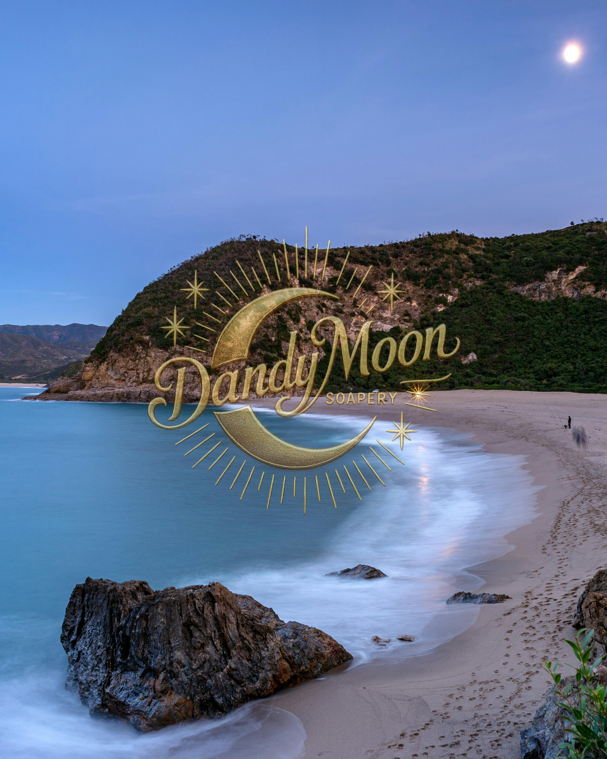 A scenic coastal beach at dusk with turquoise water, sandy shore, dark rocks, and a hillside covered in greenery under a blue sky with a visible moon. A large gold crescent moon and star graphic store logo with the text 'Dandy Moon Soapery' is overlaid across the center.