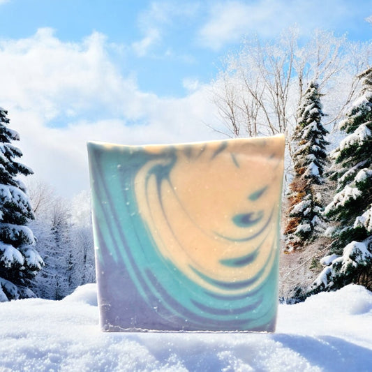 A bar of Dandy Moon Soapery's Winter Wonderland soap is displayed in a snowy outdoors setting with frosted evergreen trees and a bright blue sky in the background. The soap features a swirl design in cool tones of blue, white, and soft lavender, evoking the beauty of a winter landscape. The gentle colors and intricate patterns capture the serene and refreshing feel of a snowy day. 