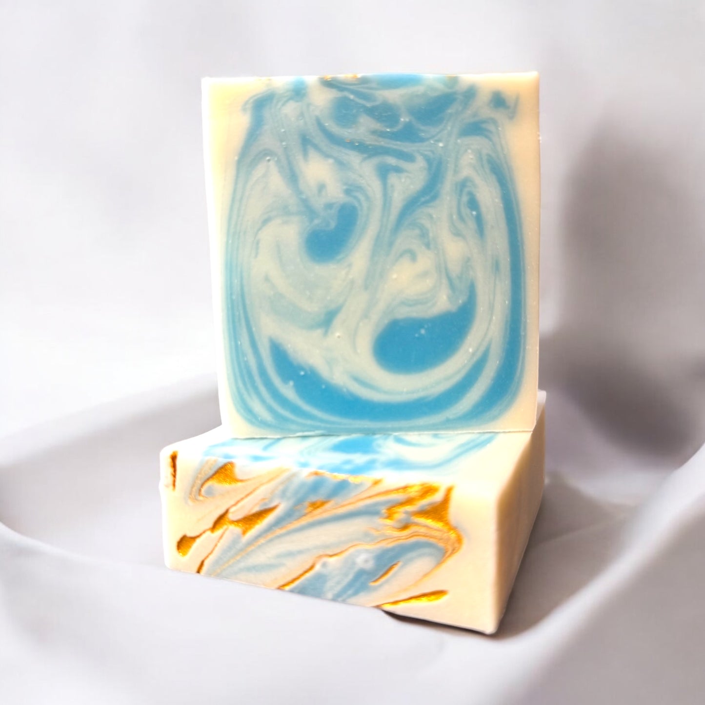 a bar of artisan soap with a vibrant swirl design in blue and white, set against a white background on a white surface. The soap has a top design of swirls of blue and white and a splash of gold. The intricate marbling and bright colors capture a sense of tranquility and natural beauty, perfect for a refreshing and uplifting self care experience. 