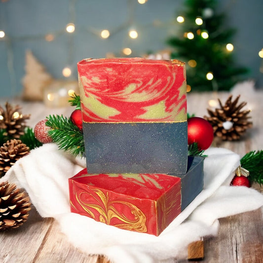 A bar of Dandy Moon Soapery's Sweater Weather soap is displayed in a festive holiday setting, surrounded by pinecones, ornaments, and evergreen branches. The soap features a cozy winter inspired red and green swirled top, and a navy blue solid bottom separated by a gold mica line. The intricate marbling and and gold accents evoke the charm of the holiday season. 