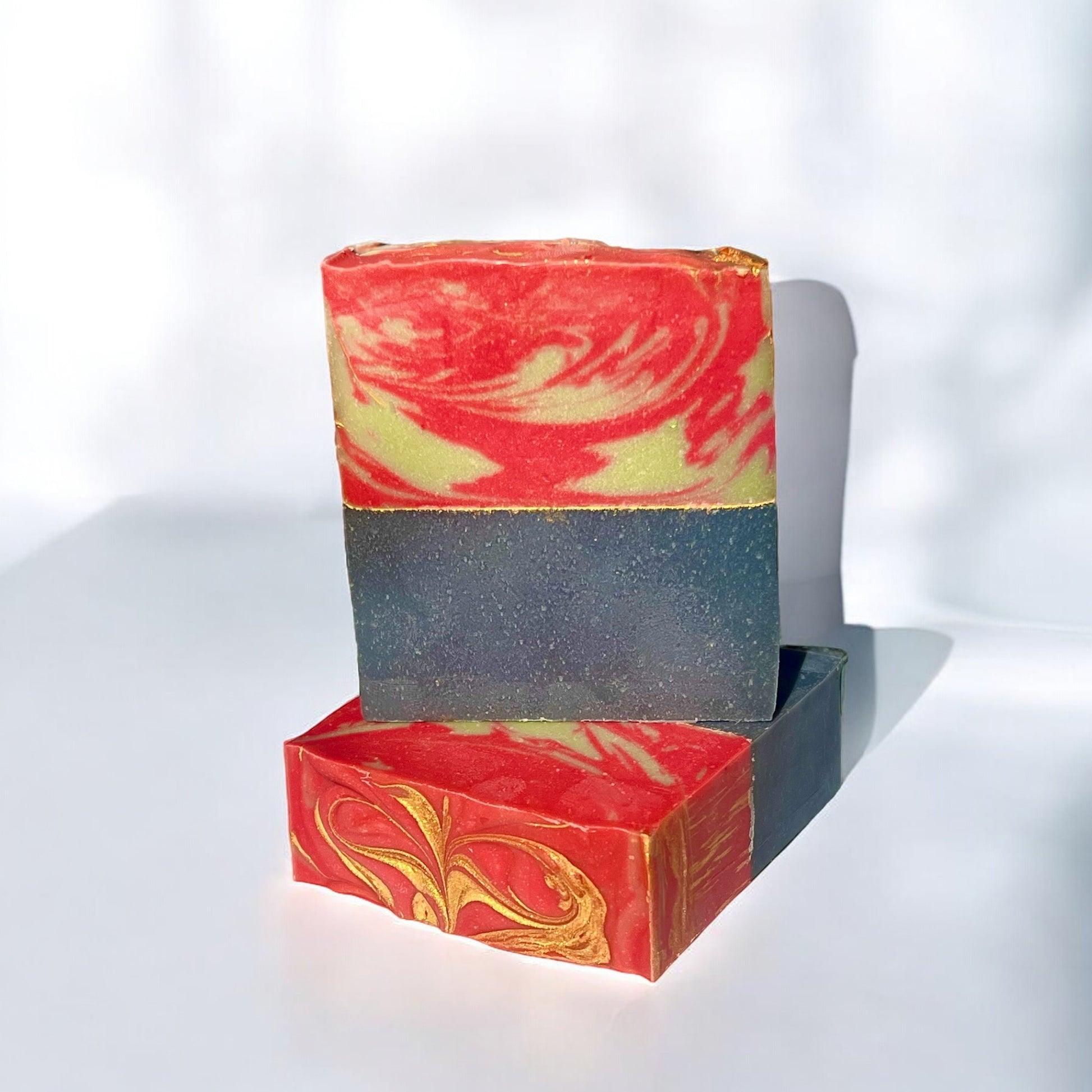 A bar of Dandy Moon Soapery's Sweater Weather soap is displayed on a white surface against a white background. The soap features a cozy winter inspired red and green swirled top, and a navy blue solid bottom separated by a gold mica line. The intricate marbling and and gold accents evoke the charm of the holiday season. 